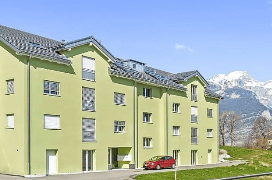 Apartment building, multiple floors, solar panels on the roof, several windows, red car parked in front, green grass, trees, mountains in the background