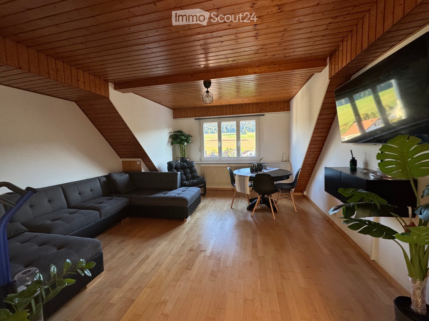 modern living room with wooden floor, gray couch, dining table, and window with green view