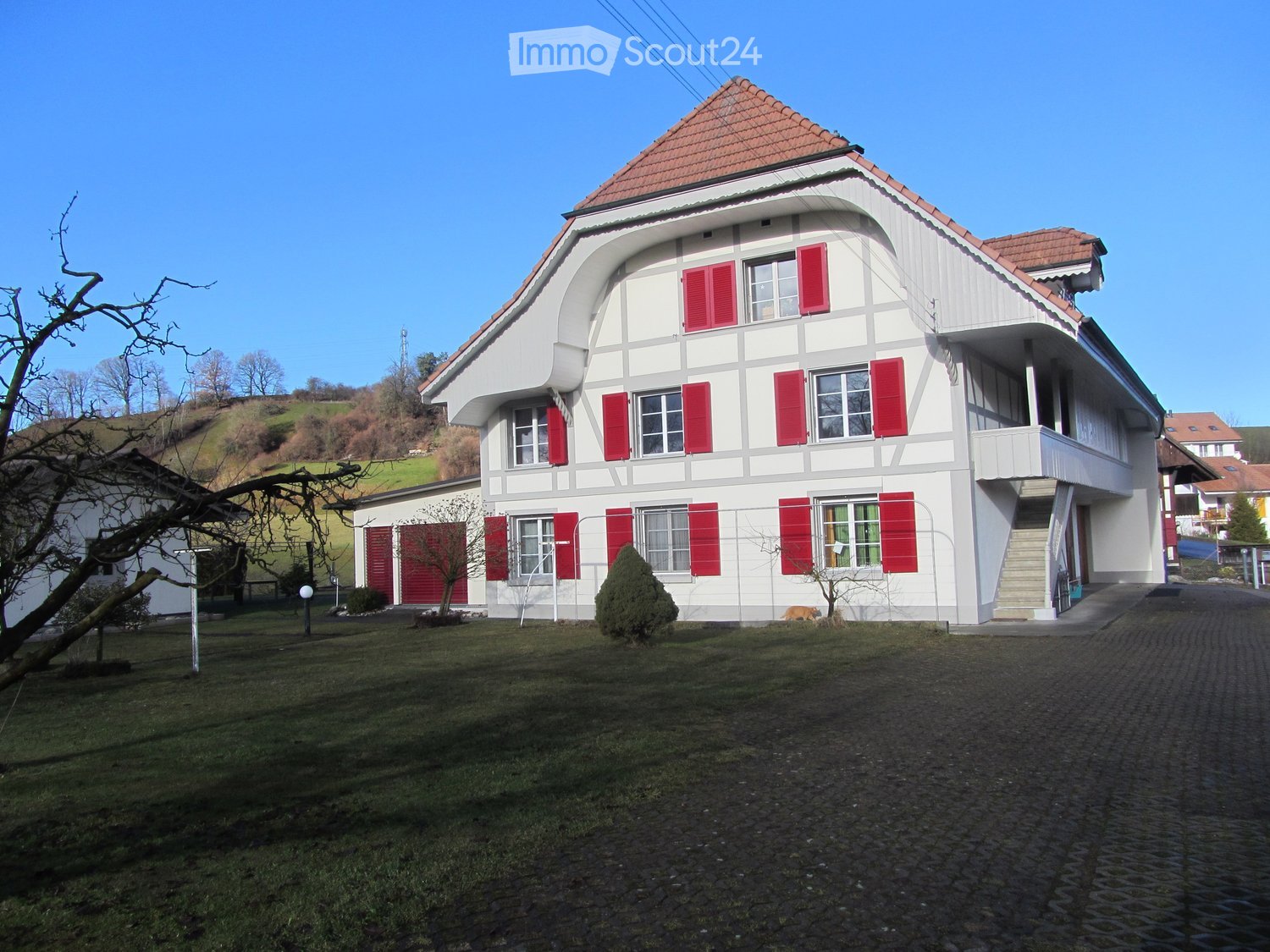 2 story house, white walls, red shutters, tiled roof, chimney, balcony, paved driveway, garden with trees and shrubs