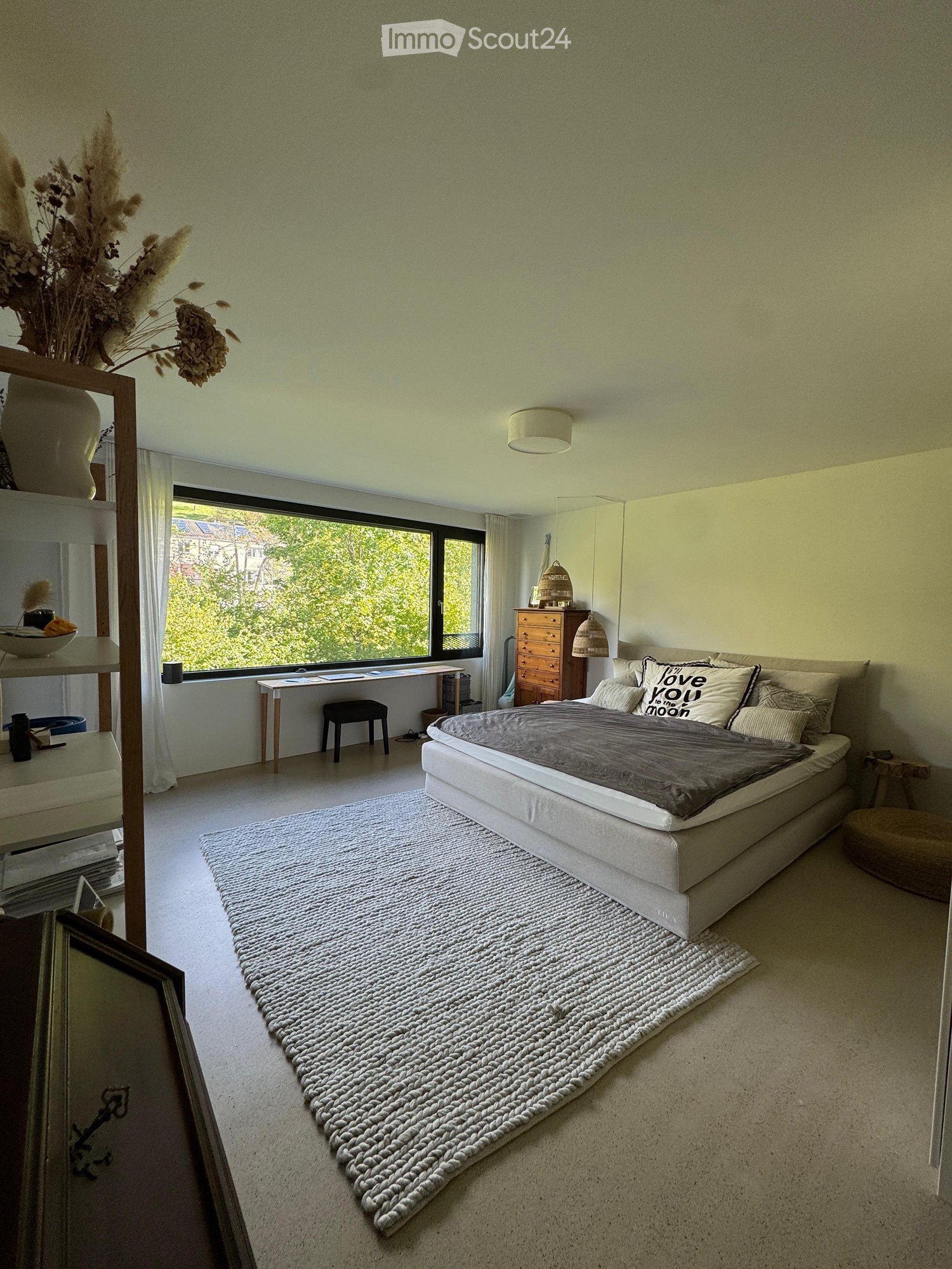 a bedroom with large windows, bed, rugs, wooden cabinet, shelves, desk, chair, pillow with text, vases