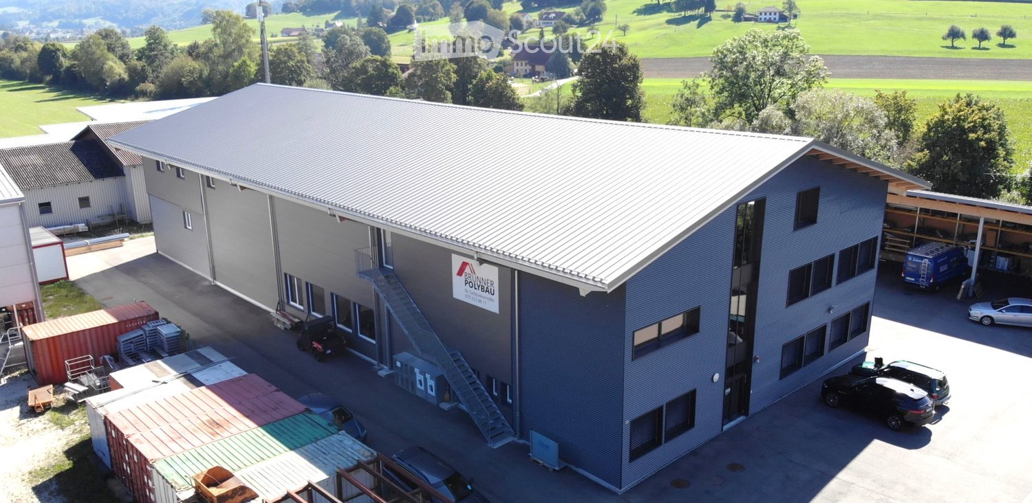 Industrial building with gray exterior, metal roof, glass windows, multiple vehicles parked in front, stairs leading up to entrance, surrounded by trees and green landscape