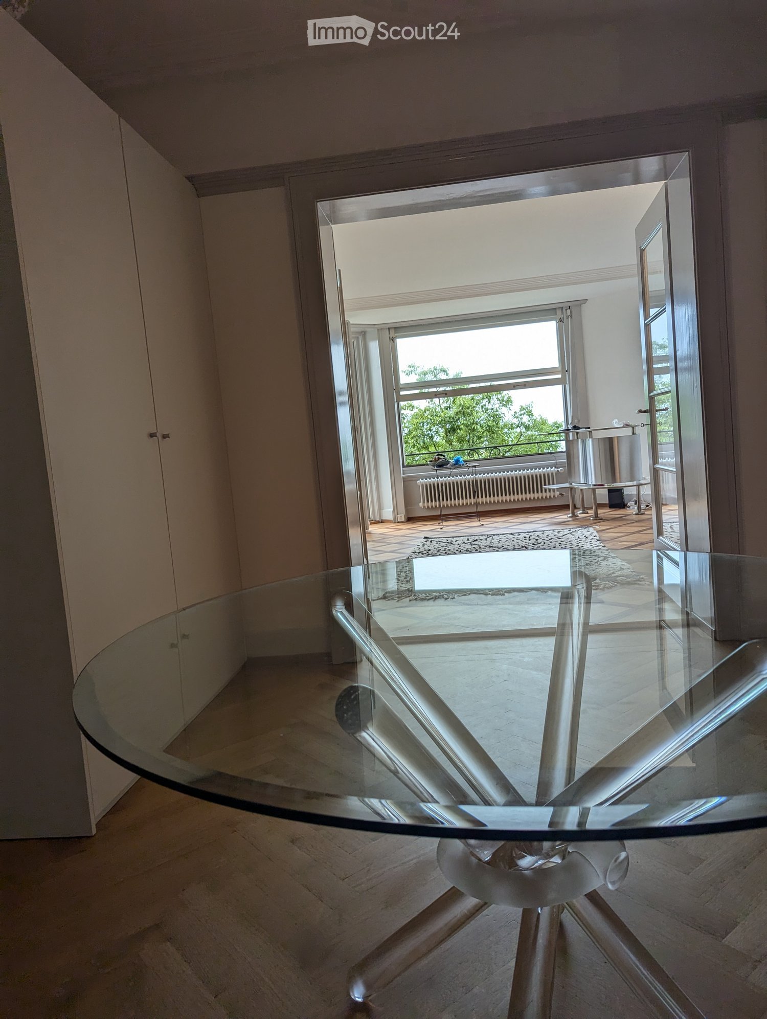 A circular glass table with white base in a room with tiled flooring, white cabinets and an open doorway leading to another room with a glass window, and a glimpse of a balcony outside
