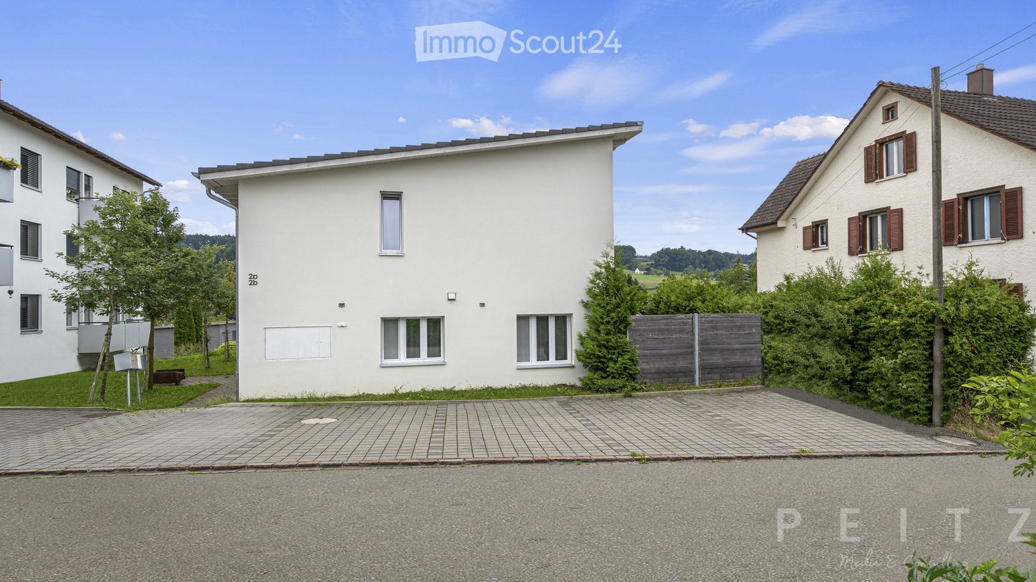 2-story white house with a flat roof, surrounded by trees and greenery, with a paved driveway or parking area in the front
