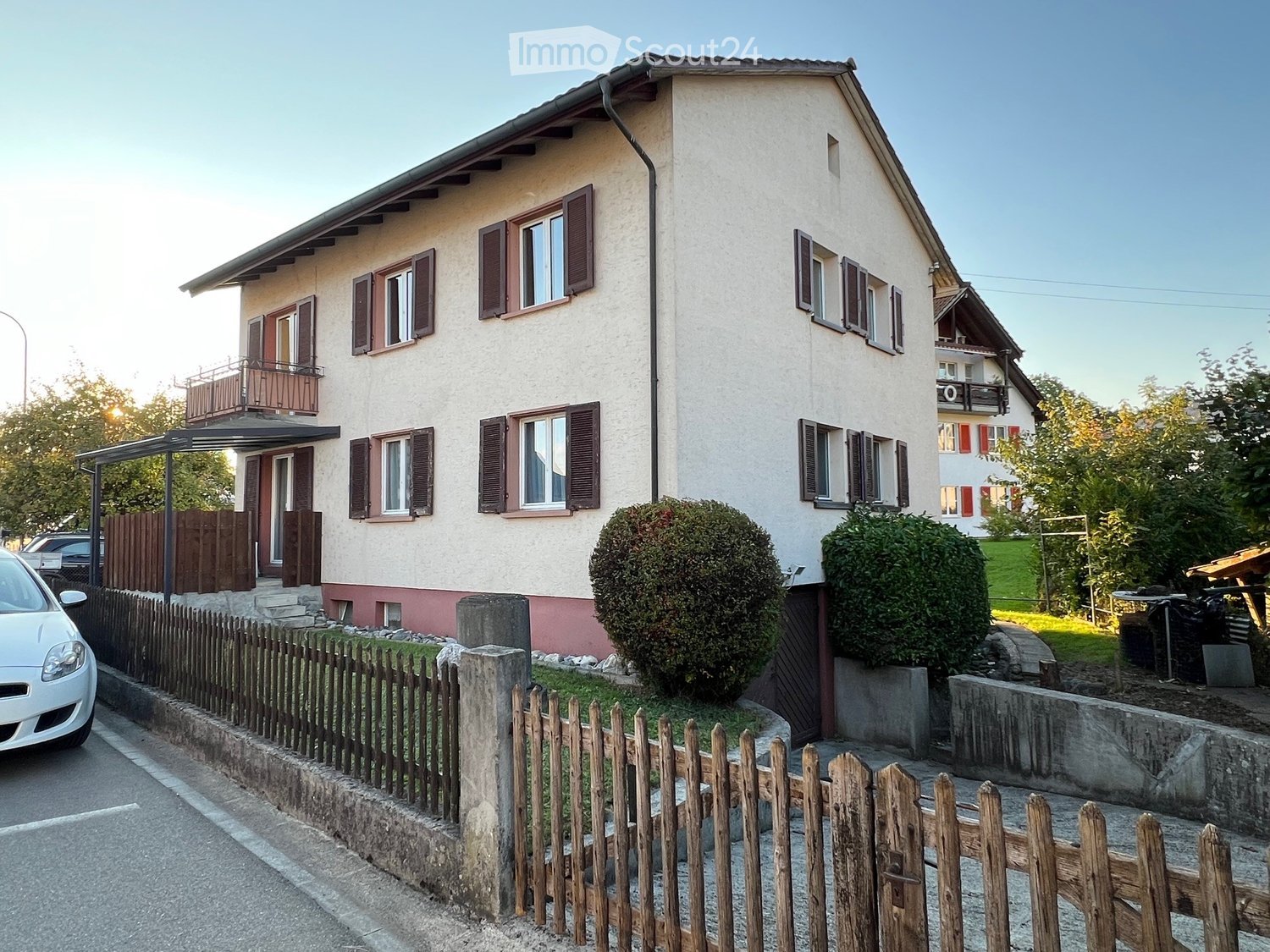 2 story house, white paint, brown windows, brown roof, fenced front yard, balcony, car parked in front