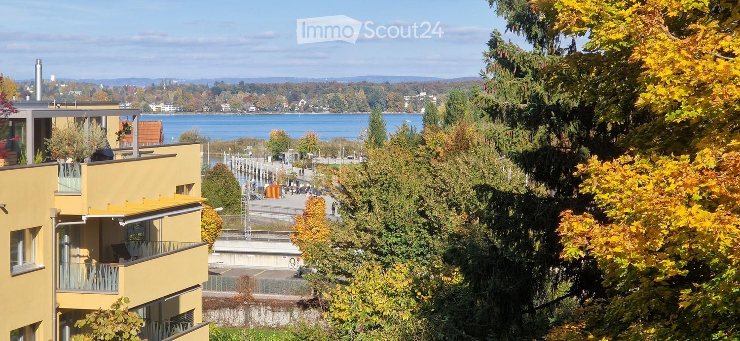 Two-story building with yellow exterior, balcony with railings, overlooking a lake and distant landscape with trees and buildings