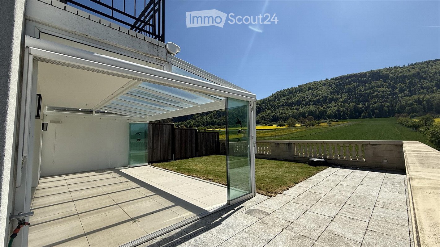 Outdoor terrace with glass wall, metal railings, concrete flooring, greenery, mountain range