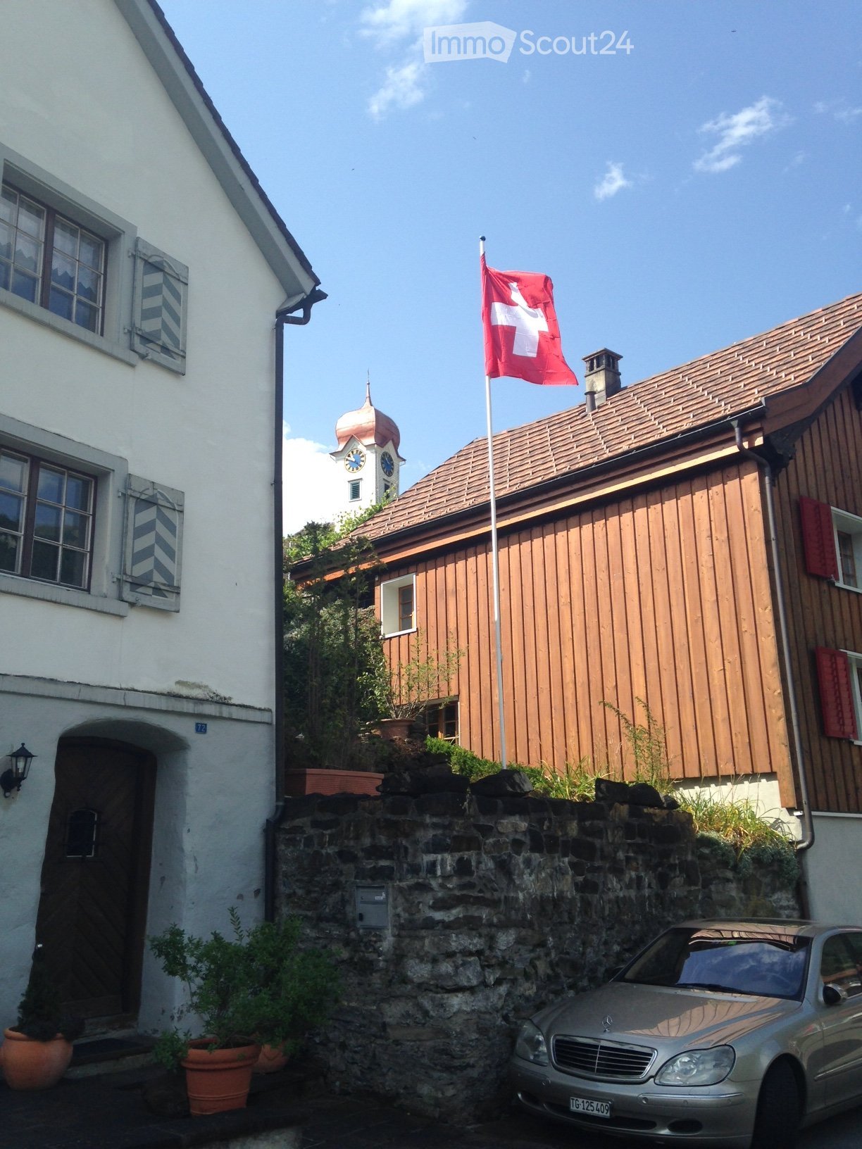 The building is painted white with brown shutters. A flag of Switzerland is on the pole in front of the building. A building with a tower on top is next to it. Potted plants are in front of the stone wall. A car is parked on the road.