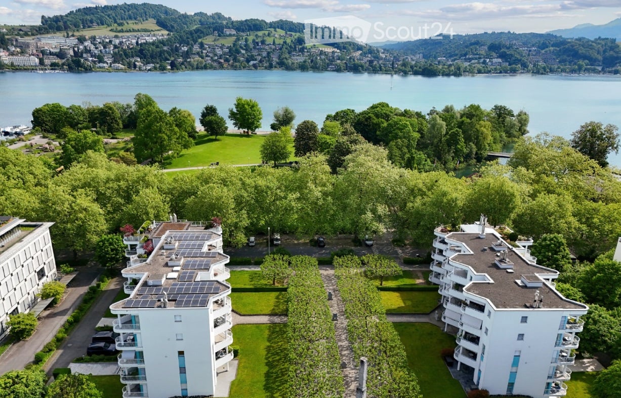 White apartment buildings, large windows, balconies, solar panels, green gardens, parking area, lake view, mountains