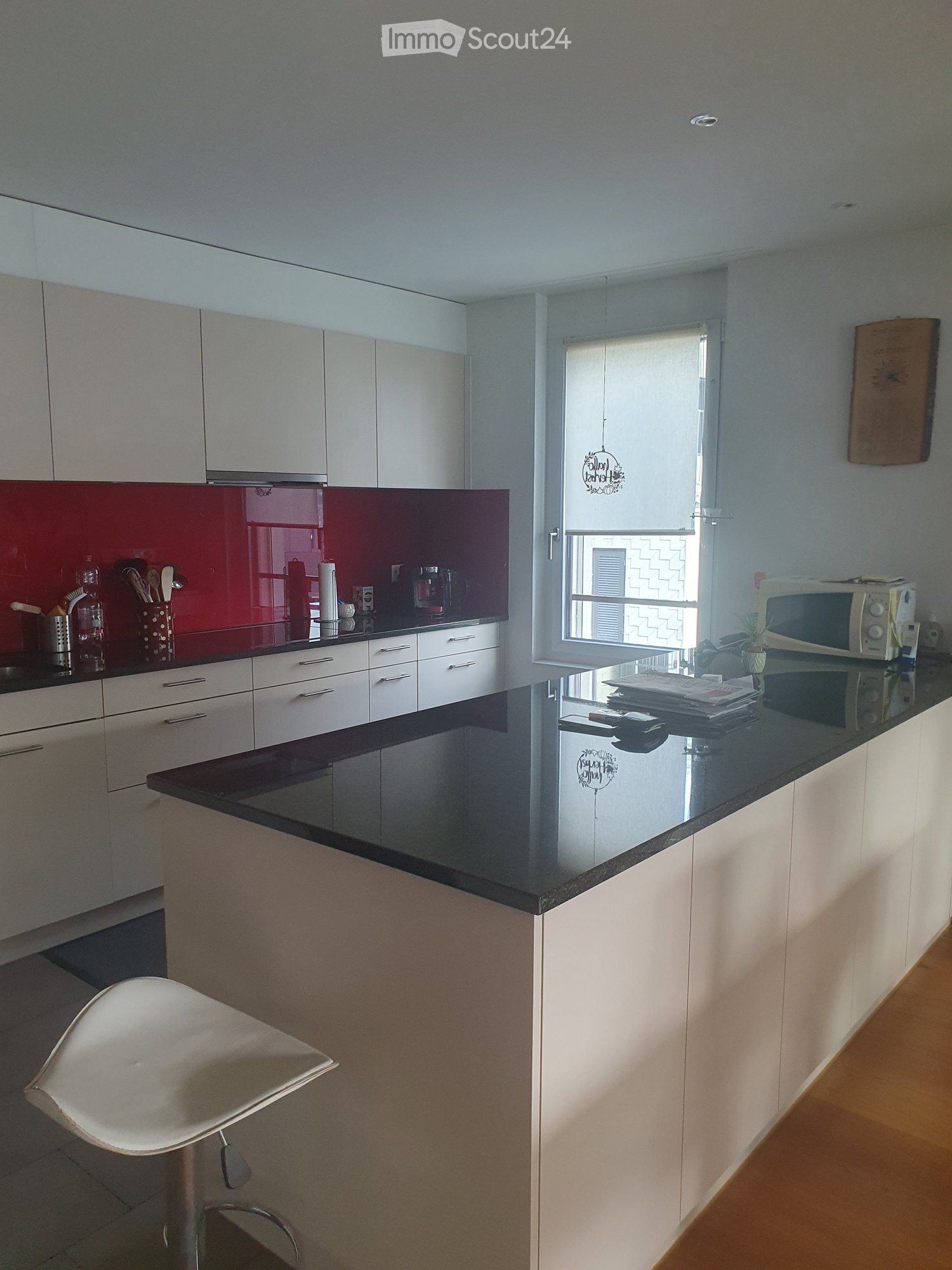 Modern kitchen with granite countertops, white cabinets, red backsplash, island, microwave, utensils, stool, and wooden floor.