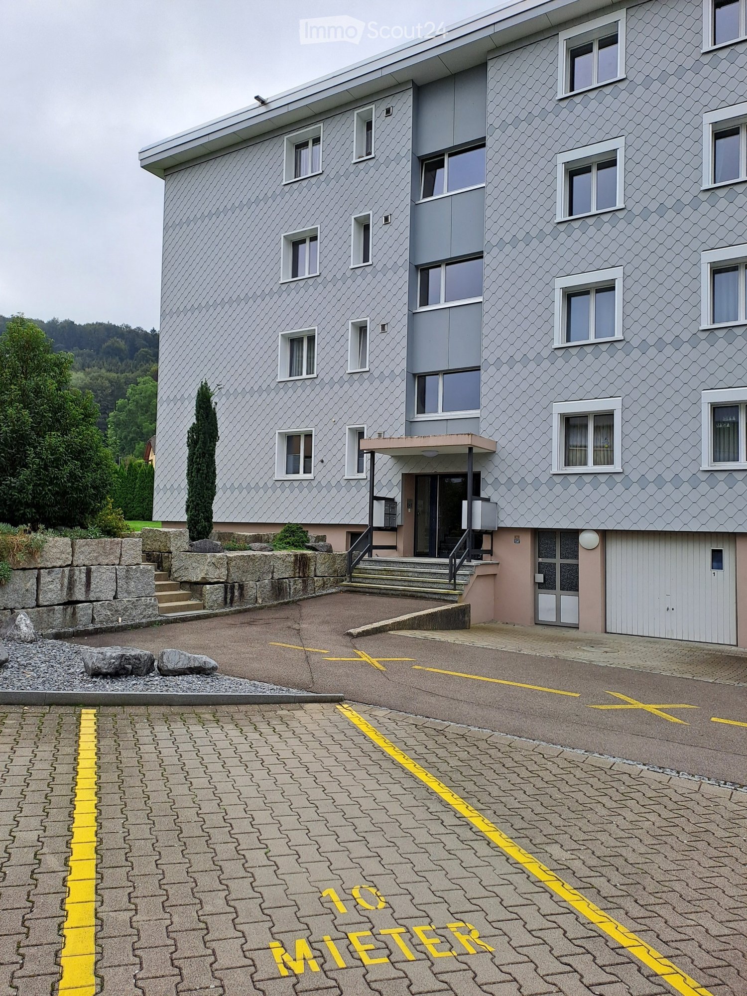 Gray building, multiple windows, garage door, steps leading up, gravel in front, yellow parking lines