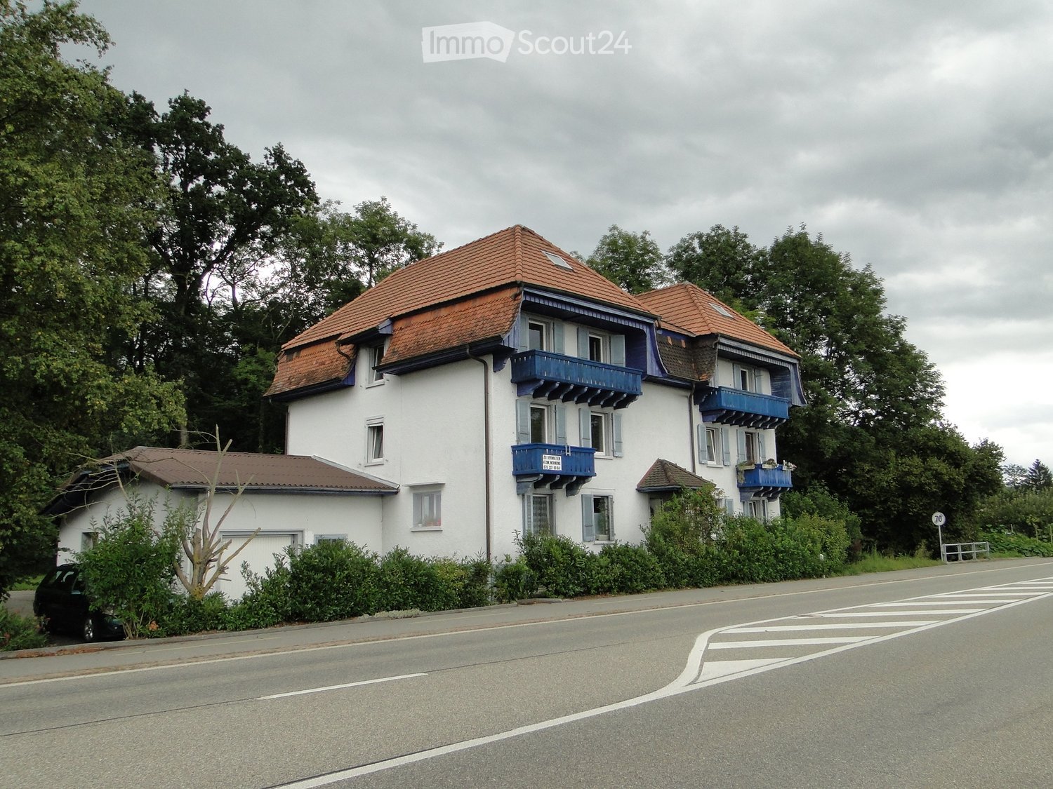 3-story apartment building with white walls, red tile roof, and blue balconies. The building is surrounded by trees and is located next to a road with a crosswalk.