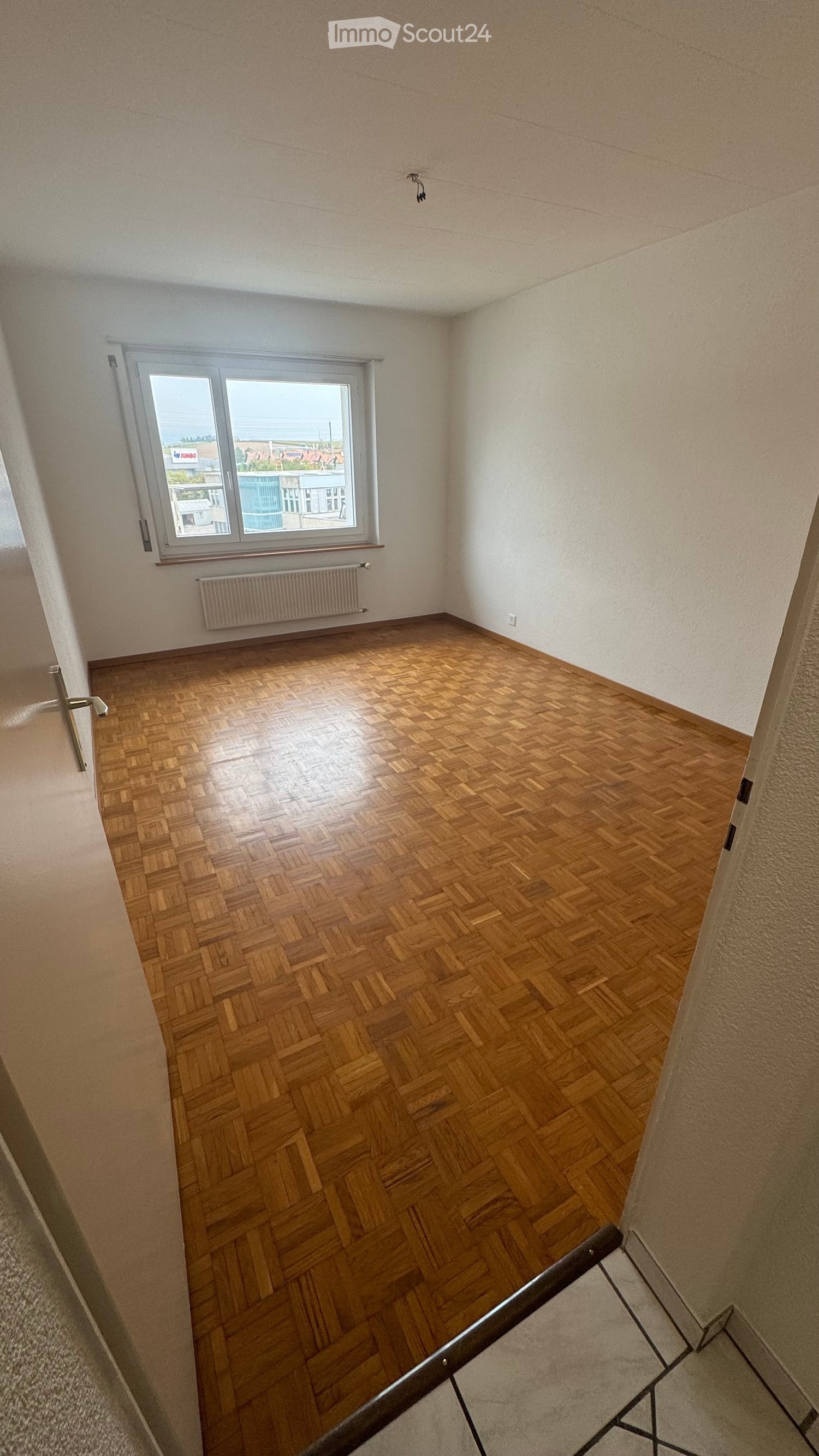 Empty room with wooden parquet flooring, large window, white walls, and a radiator.