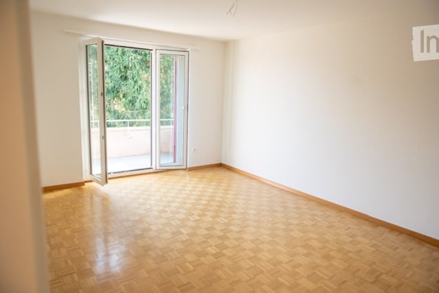Empty room with wooden floor, sliding glass doors leading to balcony, white walls.