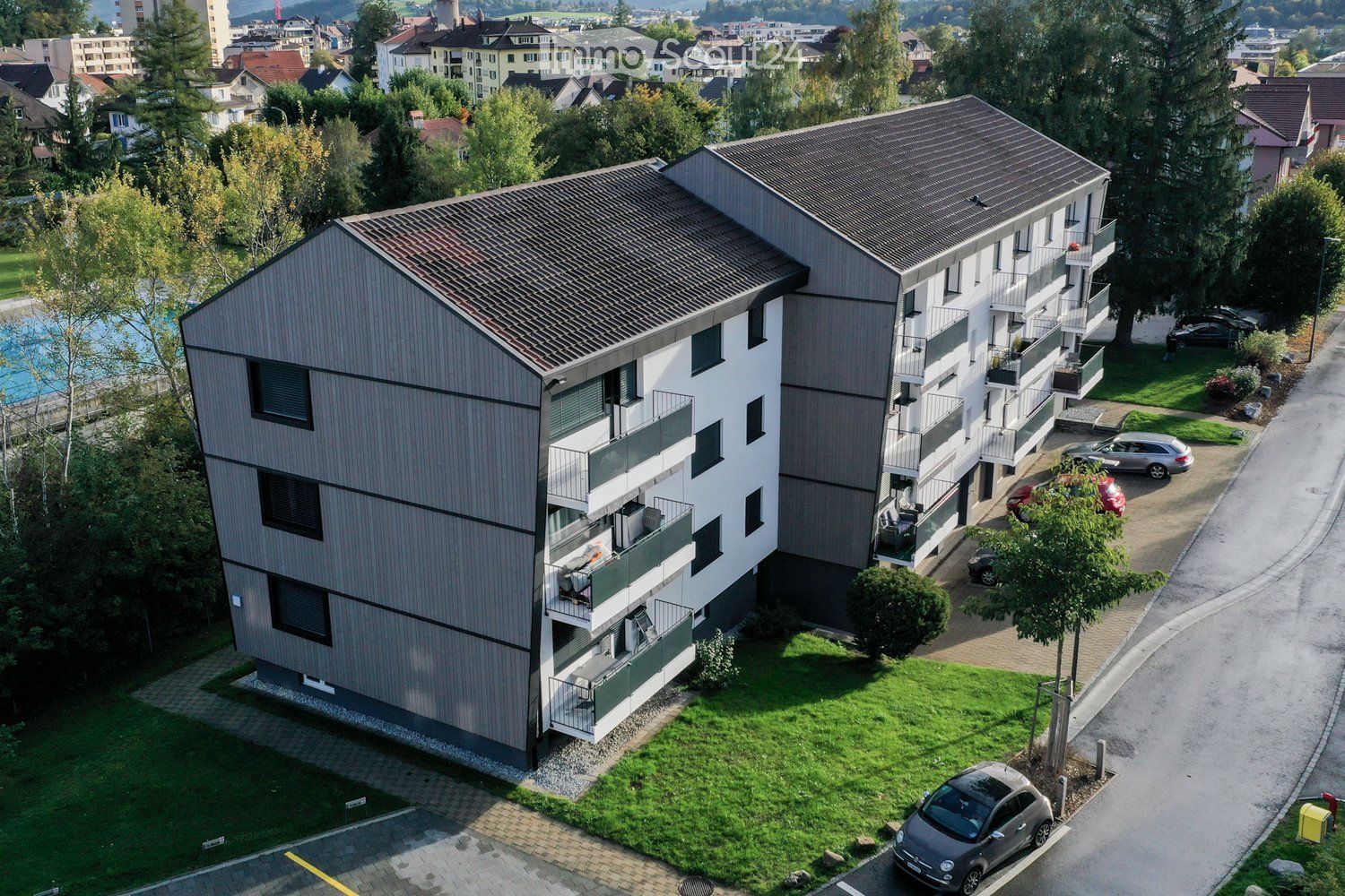 3-story building, gray exterior, many balconies, paved driveway with parking spaces, grassy area in front