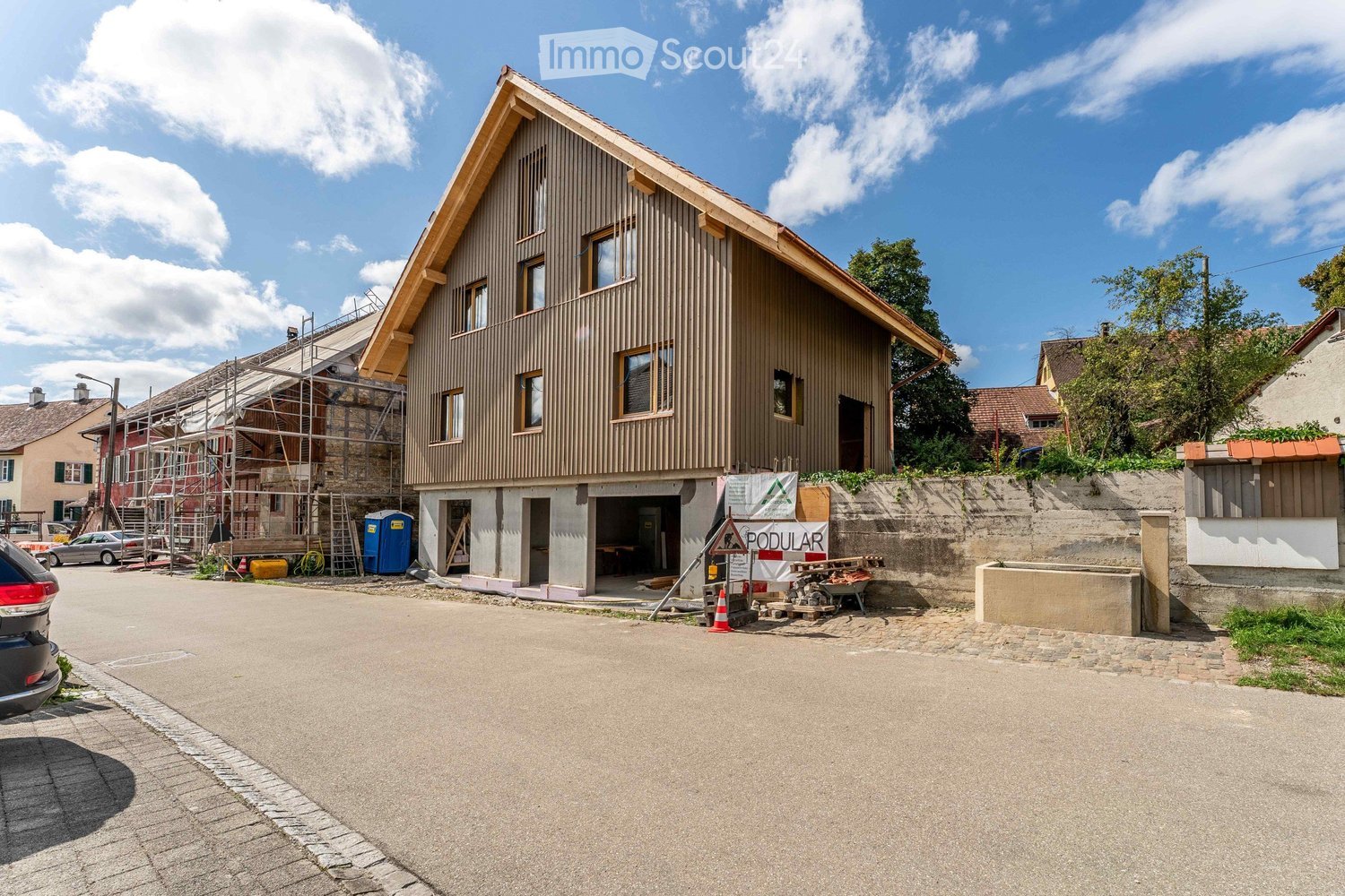 A 2-story house with a sloping roof, made of light brown walls, several windows, and an open garage. It is currently under construction, with some scaffolding around the building. There are cars parked on the side of the road and a small wall in front of