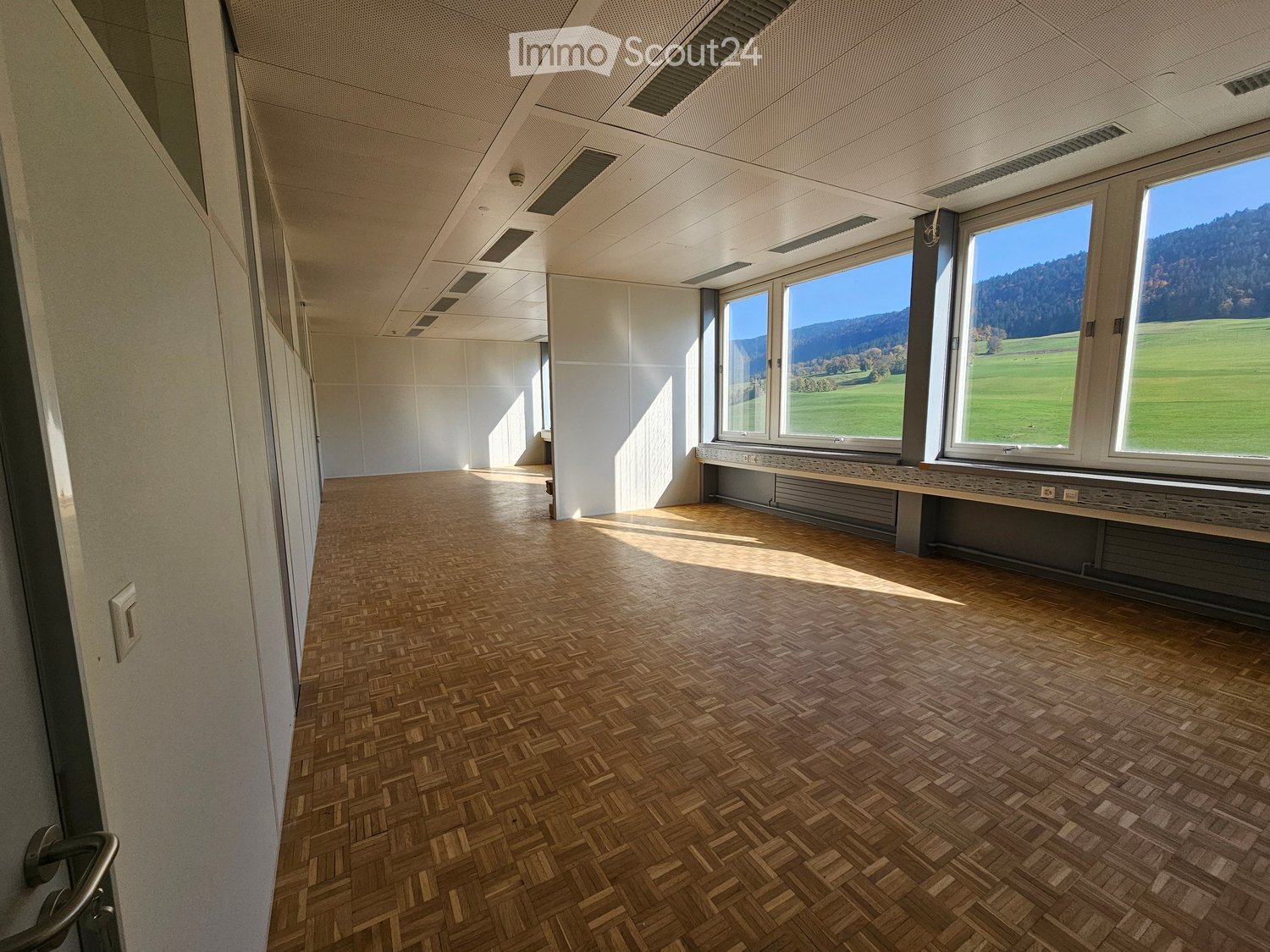 Empty room with wooden floor, white walls, windows with view of the landscape, and built-in benches along the windows.