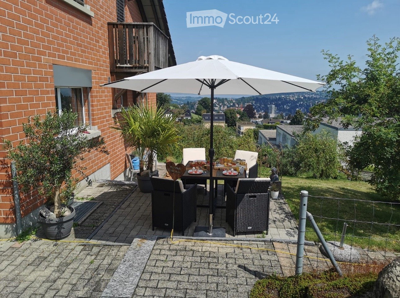 outdoor seating area with table and chairs, large umbrella, brick wall, wooden railing, potted plants, fenced area