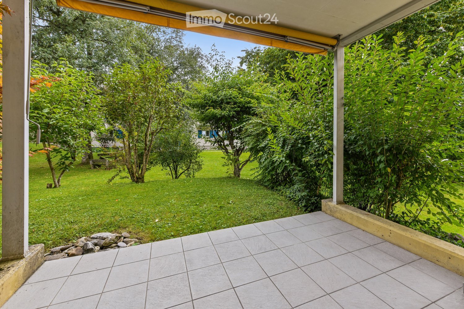 tiled terrace with yellow awning, green lawn, trees, rocks