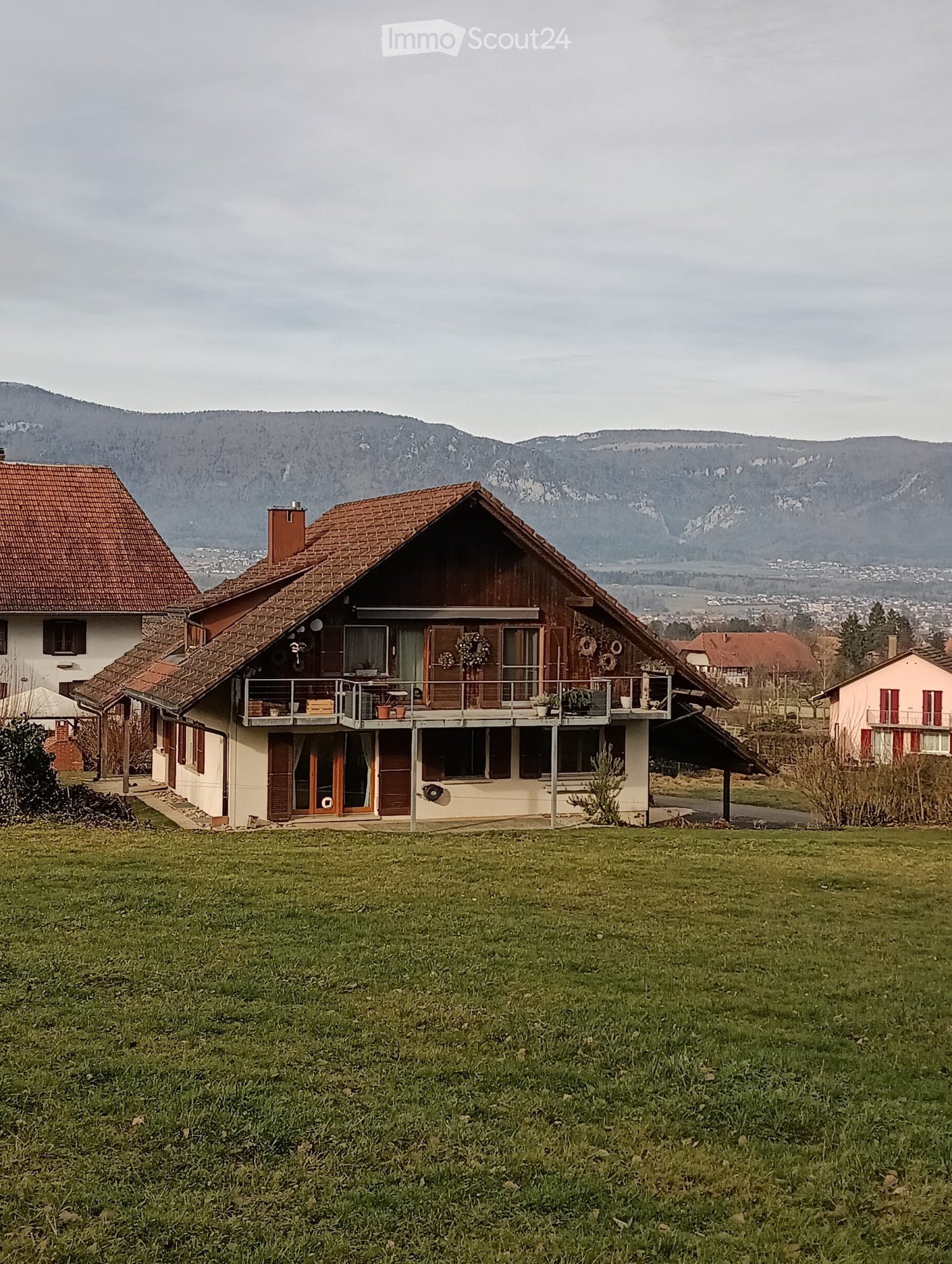 Traditional rural house, two stories, brown roof, balcony, surrounded by green field, mountains in the background