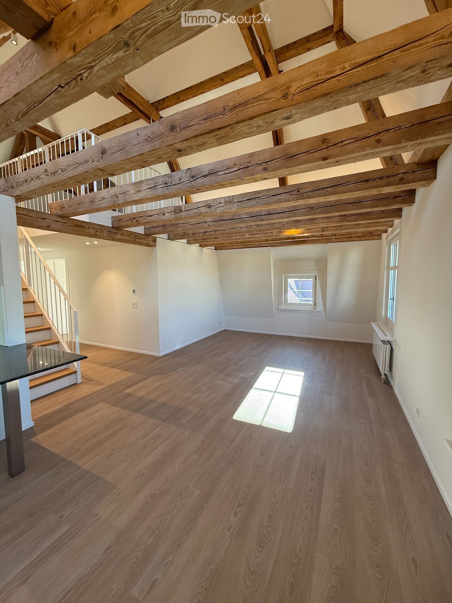 Open concept living space with exposed wooden beams on the ceiling, hardwood flooring, and a window providing natural light. The room appears to be a living area with a potential fireplace feature visible.