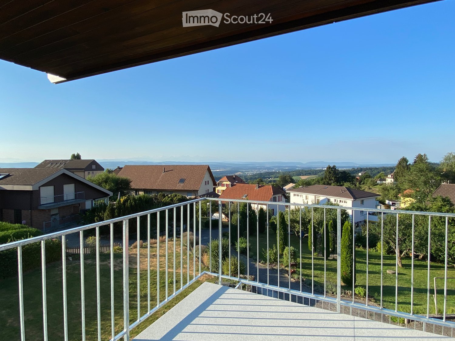 Outdoor balcony with metal railing, grass lawn, tree, and buildings in the distance.