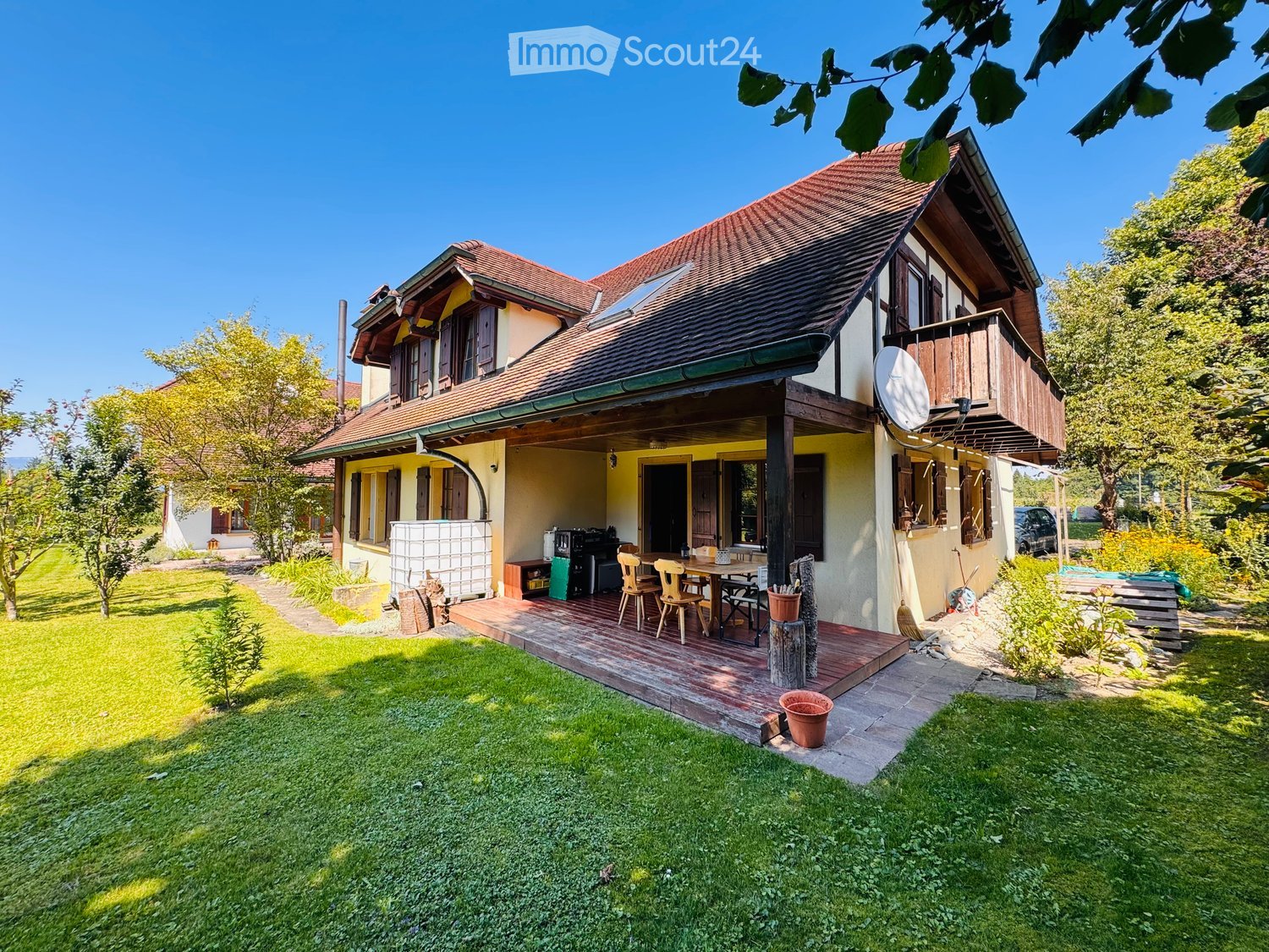 House with a wooden deck, solar panel on the roof, small balcony, patio with outdoor dining set, greenery around the house