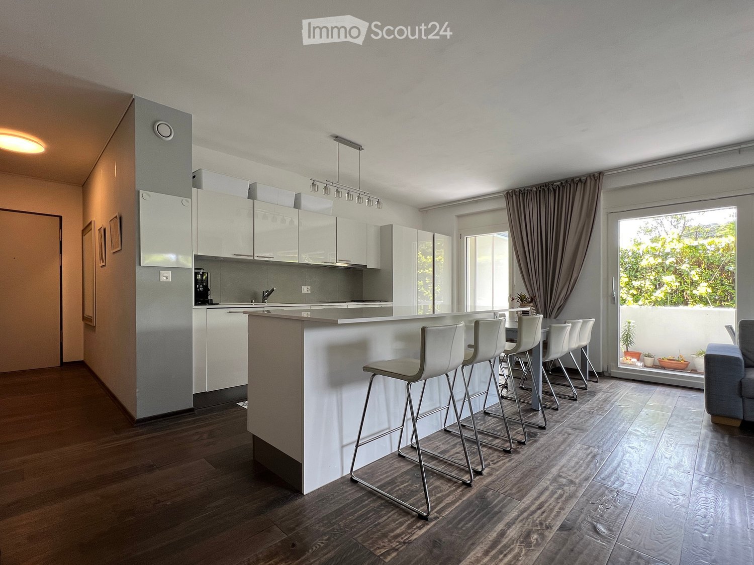 Modern kitchen with white countertops, wooden floors, open shelving, bar stools, ceiling lights, a door, and a balcony with plants.