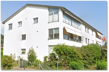 Three-story building with multiple windows, a balcony with an awning, and several plants in front.
