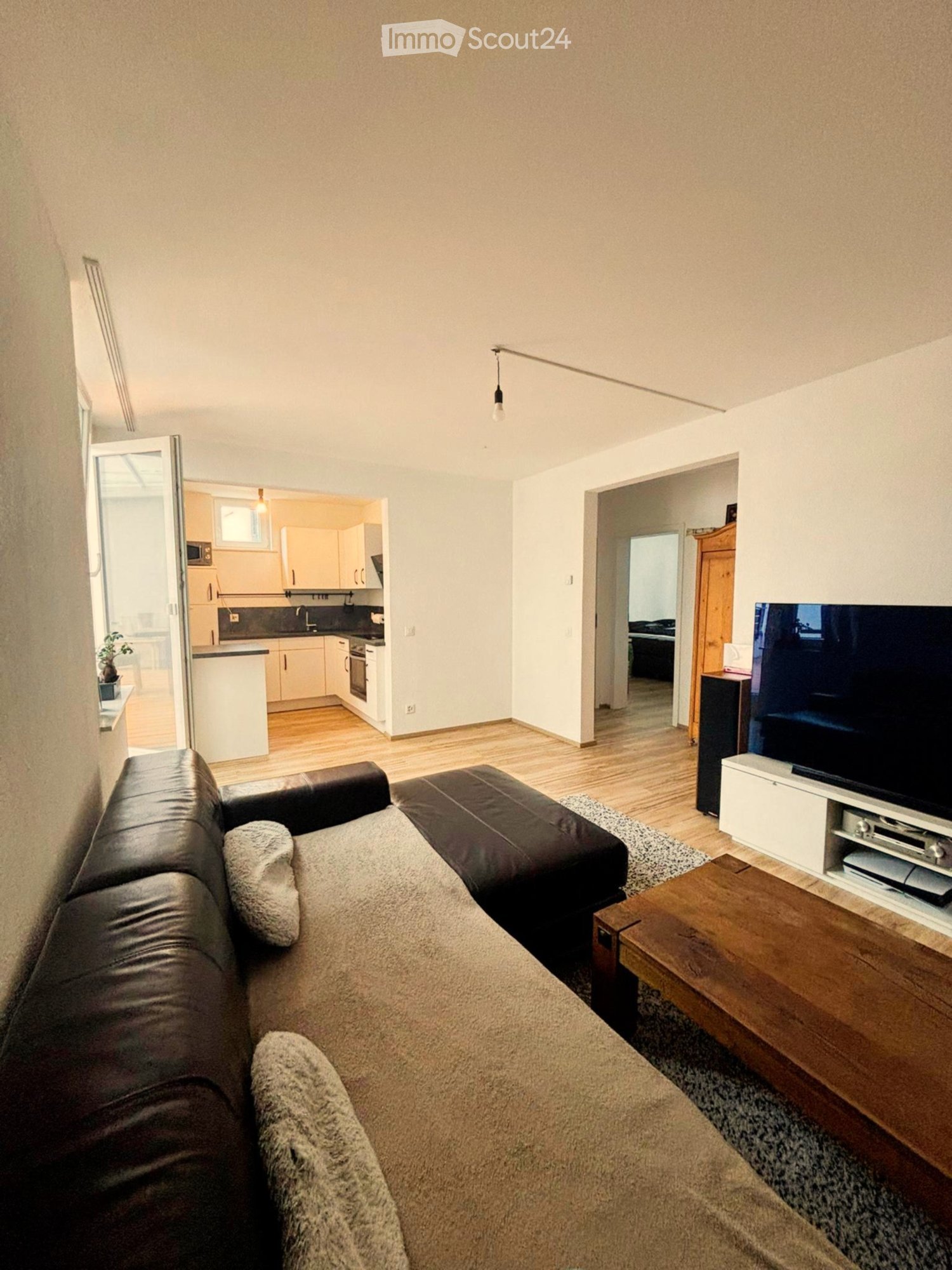 large couch, carpet, coffee table, TV, open door leading to balcony, kitchen in the background