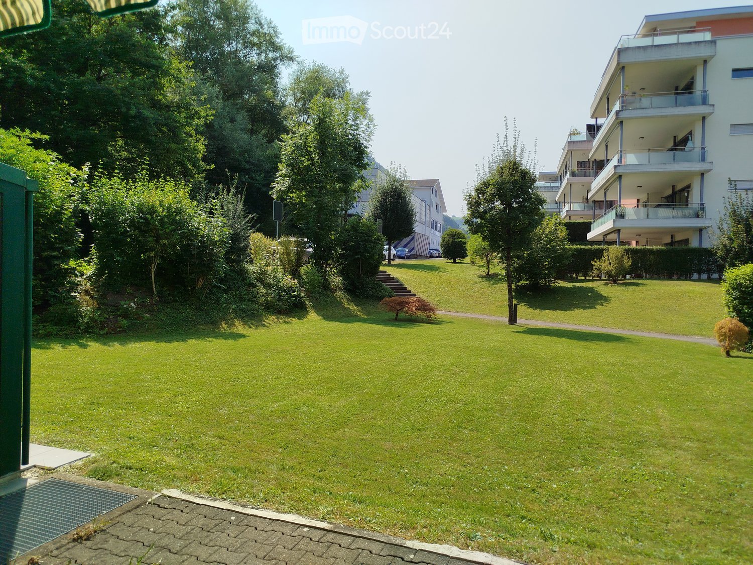 A park-like garden area with a grassy lawn, surrounded by greenery, and trees, leading to a building with balconies