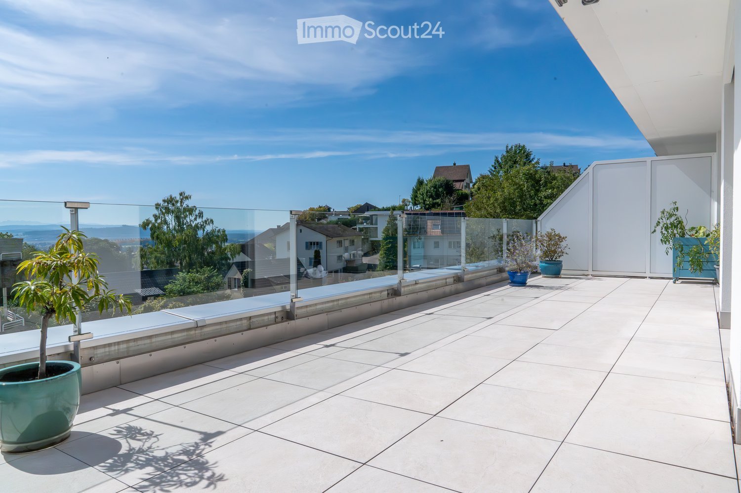 Tiled balcony floor, glass railing, plants in pots, mountain view, buildings in background