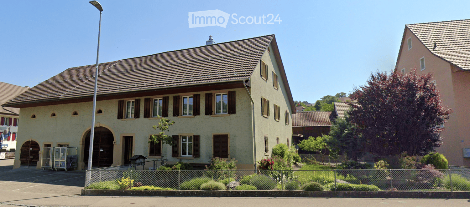 Two-story house with brown roof, beige walls, and brown shutters. Arched doorway with small archways and double doors. Garden with various plants and trees in front.