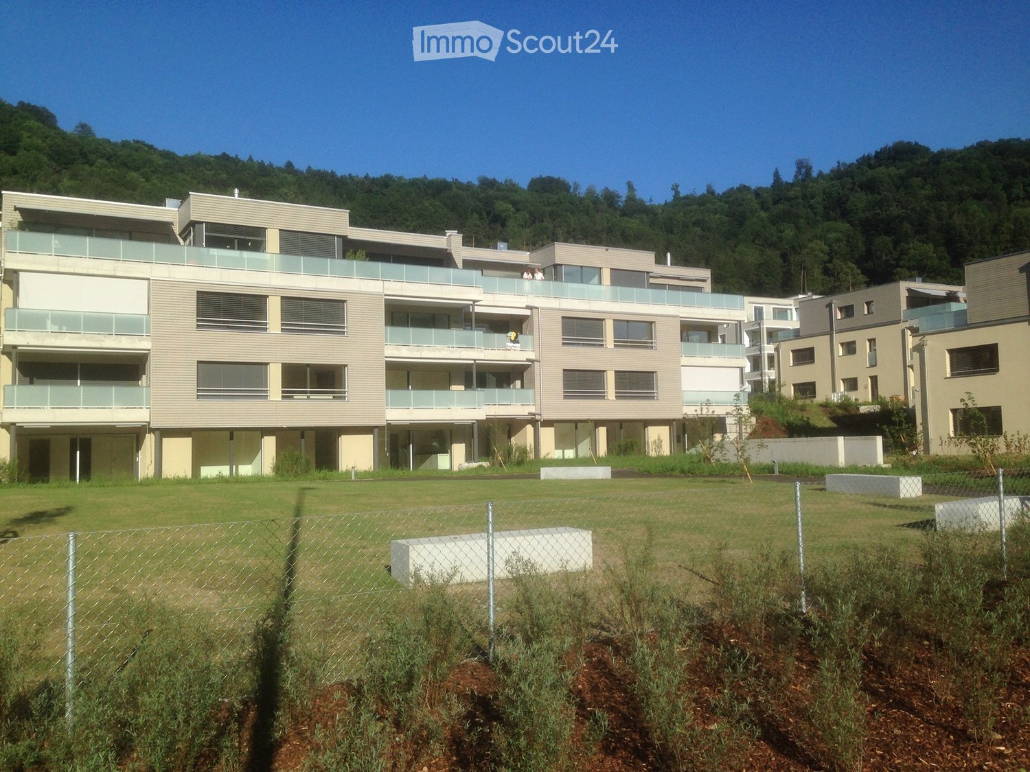 Modern apartment building with multiple levels, large balconies, and windows. It is surrounded by a large grassy field and a chain link fence. There are no visible parking facilities or other amenities.