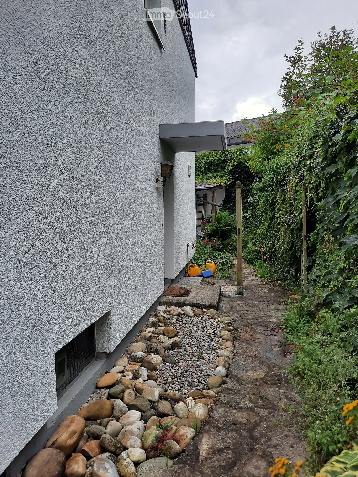 Backyard of a house with a grey wall, pathway, rocks, greenery, plants, and a wooden post