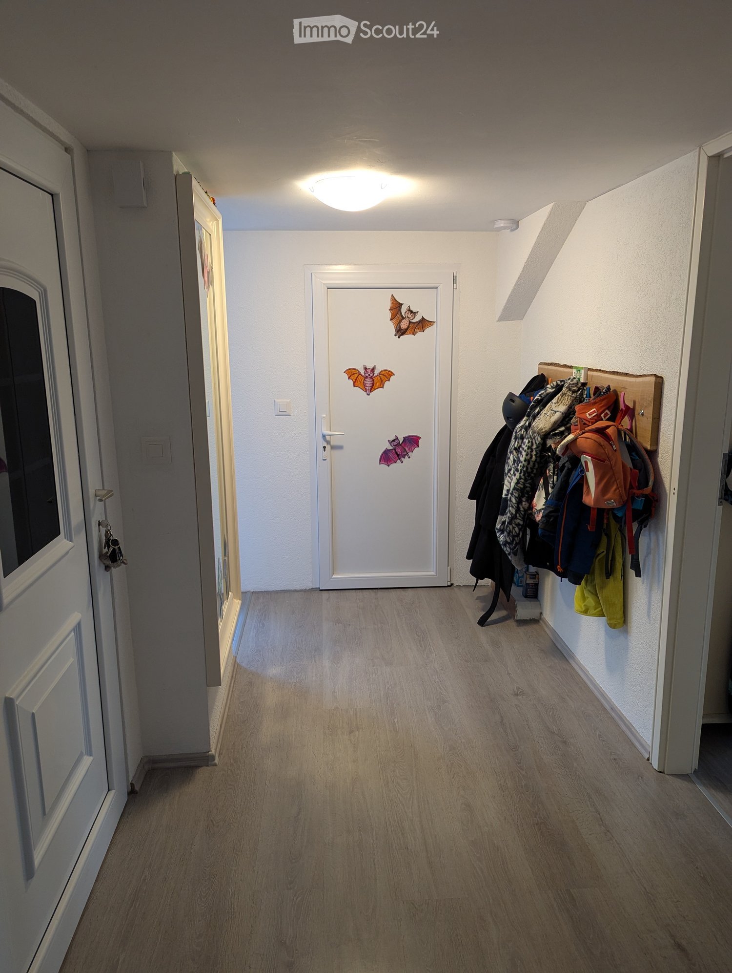 A white hallway with wooden floors and white walls, featuring a ceiling light, a door adorned with bat stickers, and clothes hanging on the wall.