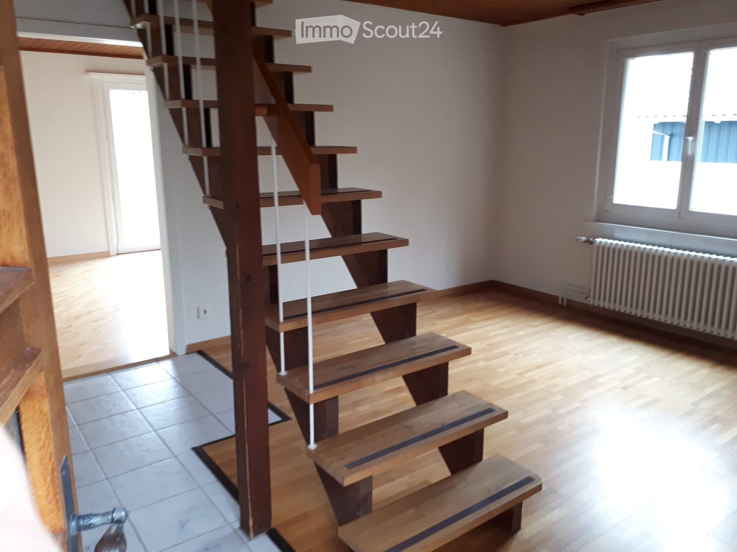 Empty room, wooden staircase with railing, white walls, wooden floor, radiator next to the window, natural light coming through the window
