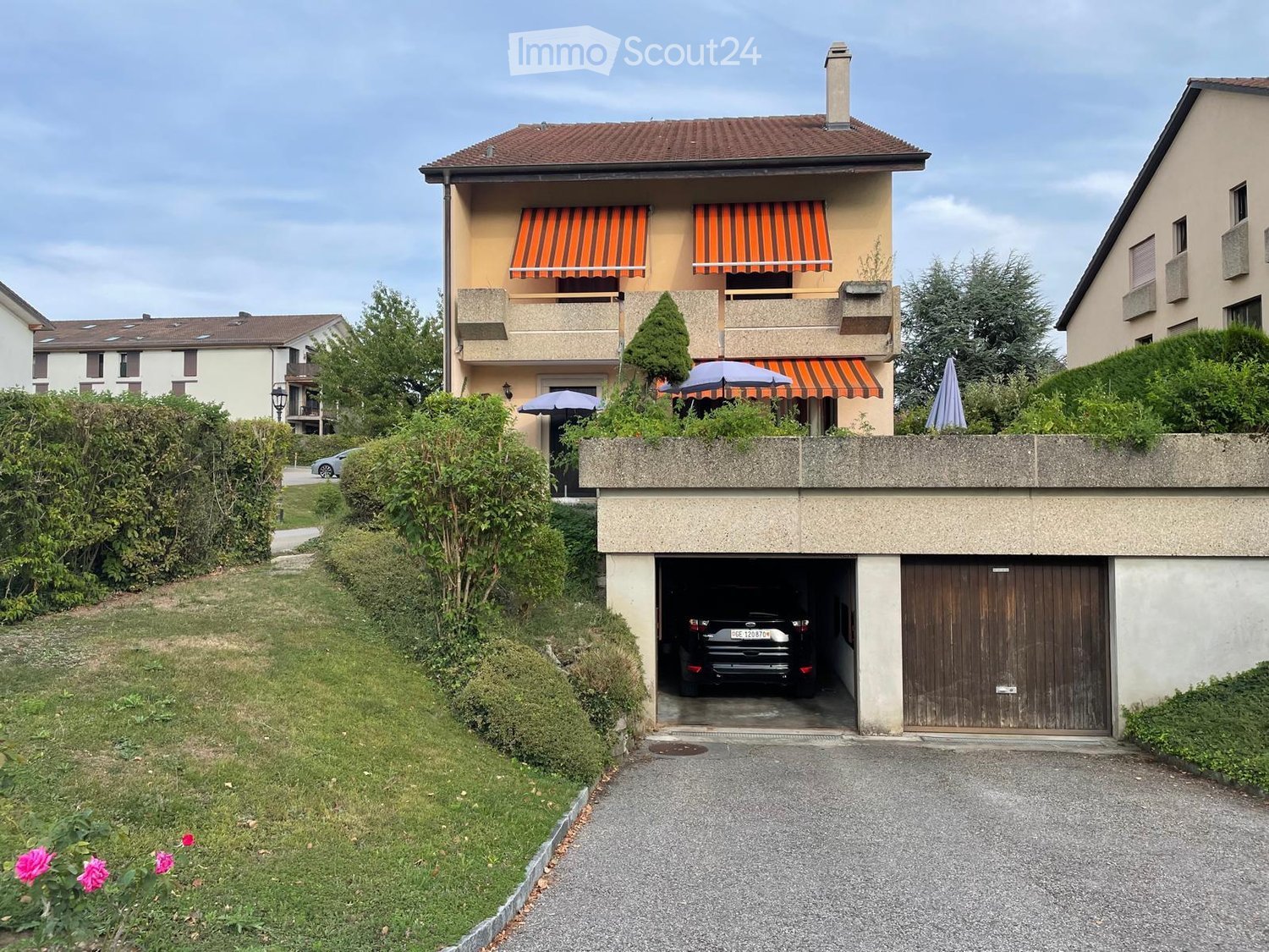 2 story apartment building, beige facade, brown roof, garage with a car inside, small garden, bushes and flowers