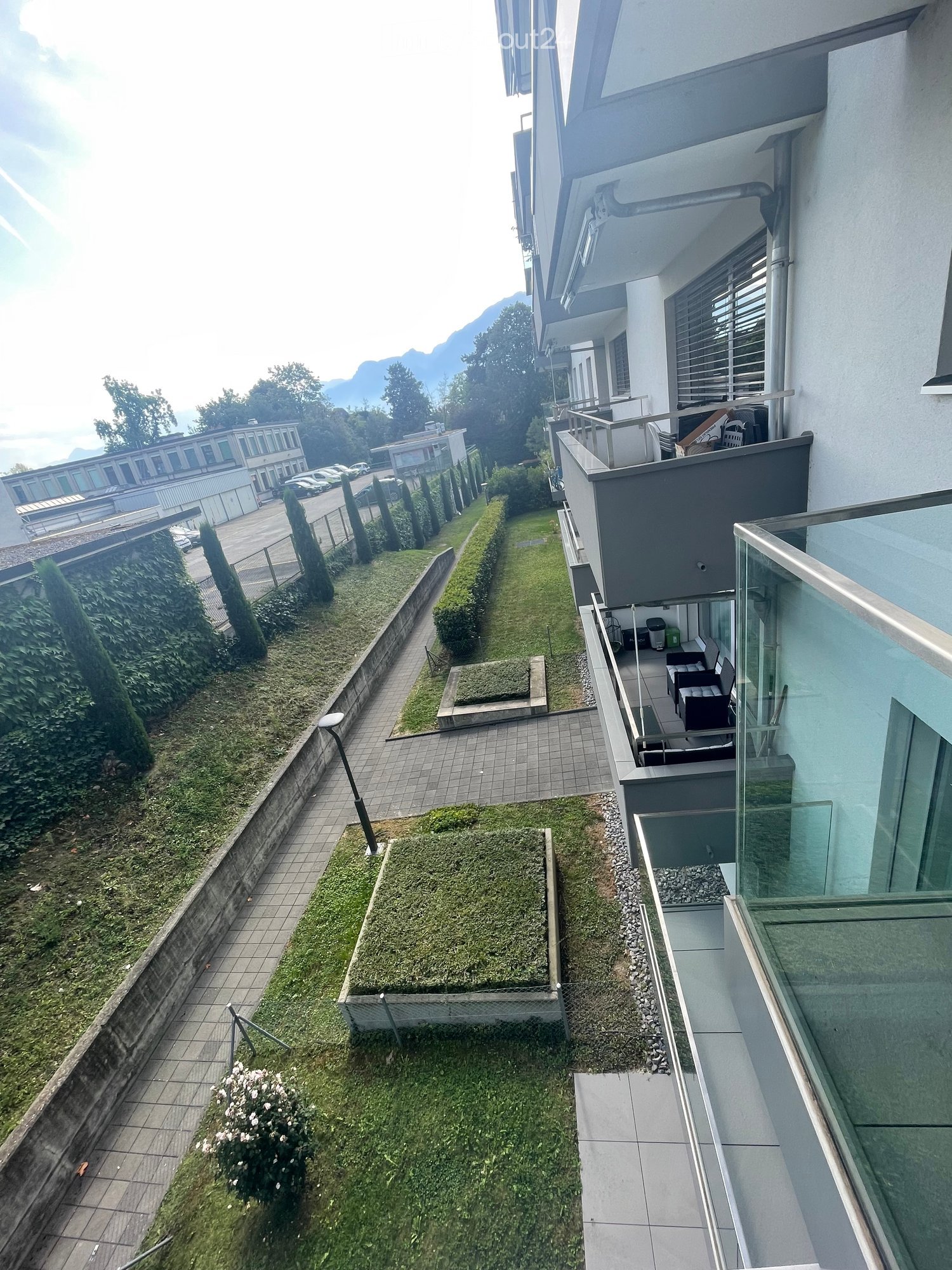 An outdoor area with multiple buildings, grass patches, pathways, trees, and mountains in the distance.