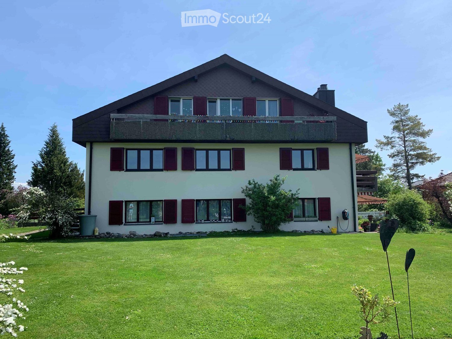 2-story house with green exterior walls, black roof, and red shutters. Large balcony on the second floor. Grassy lawn and landscaping in the front yard. Appears to have a parking area or driveway.