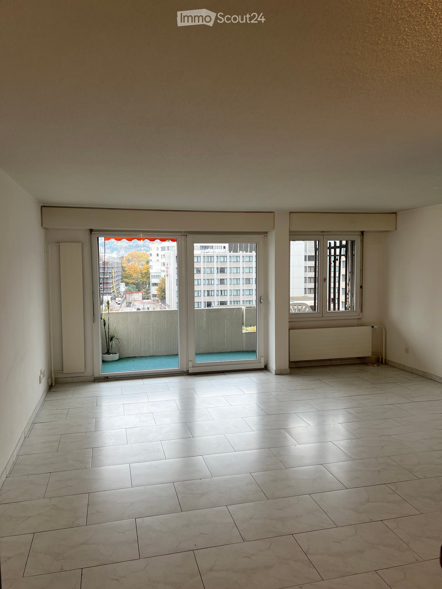 Large room with sliding glass doors opening to a balcony, white tiled floor, two windows on the right side, balcony railing