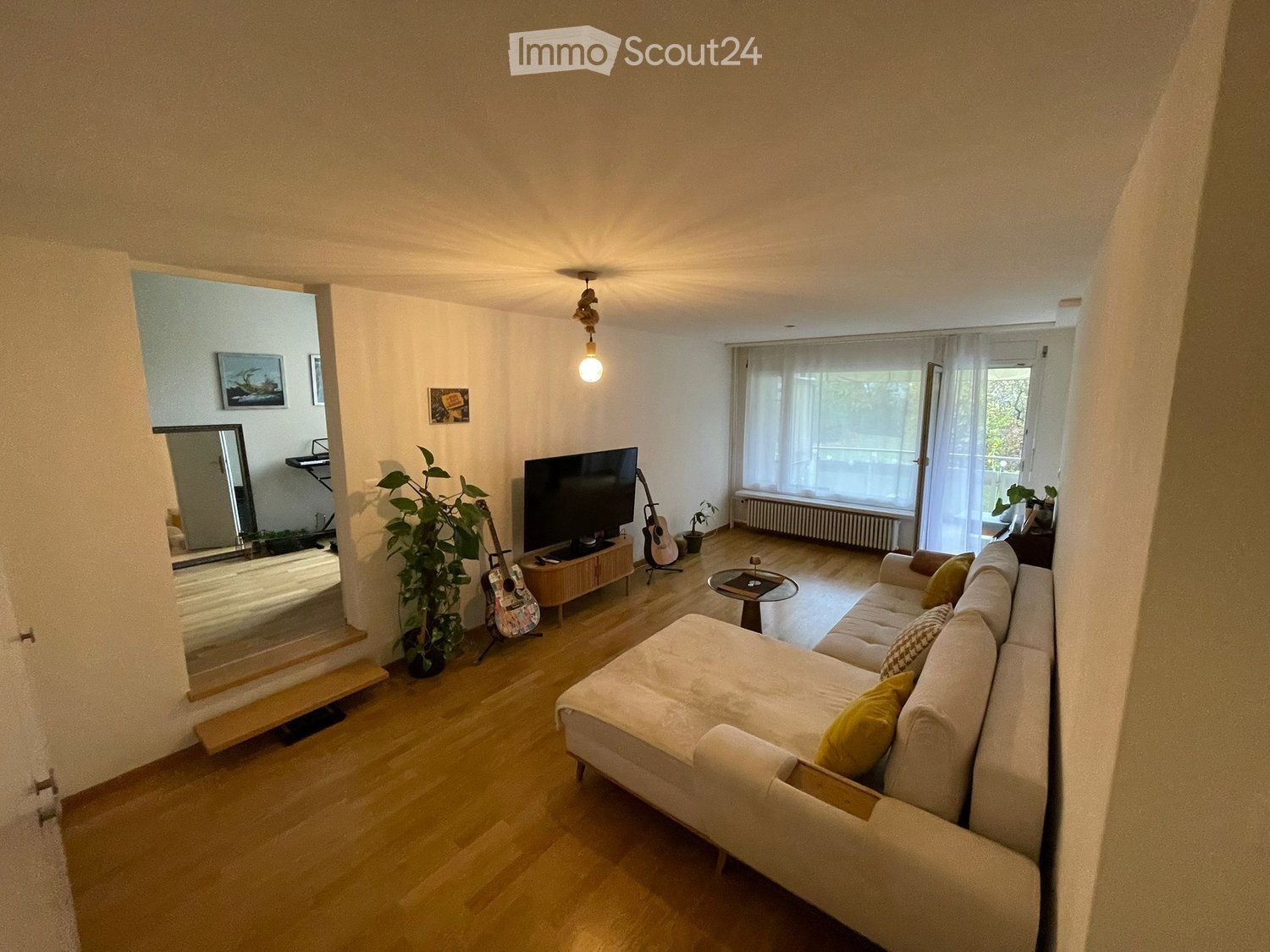 Modern, spacious living room with hardwood floors, large windows with white curtains, a white sofa with yellow cushions, a flat-screen TV, a wooden TV stand, a round table, and potted plants. It also features a radiator and two guitars leaning against the