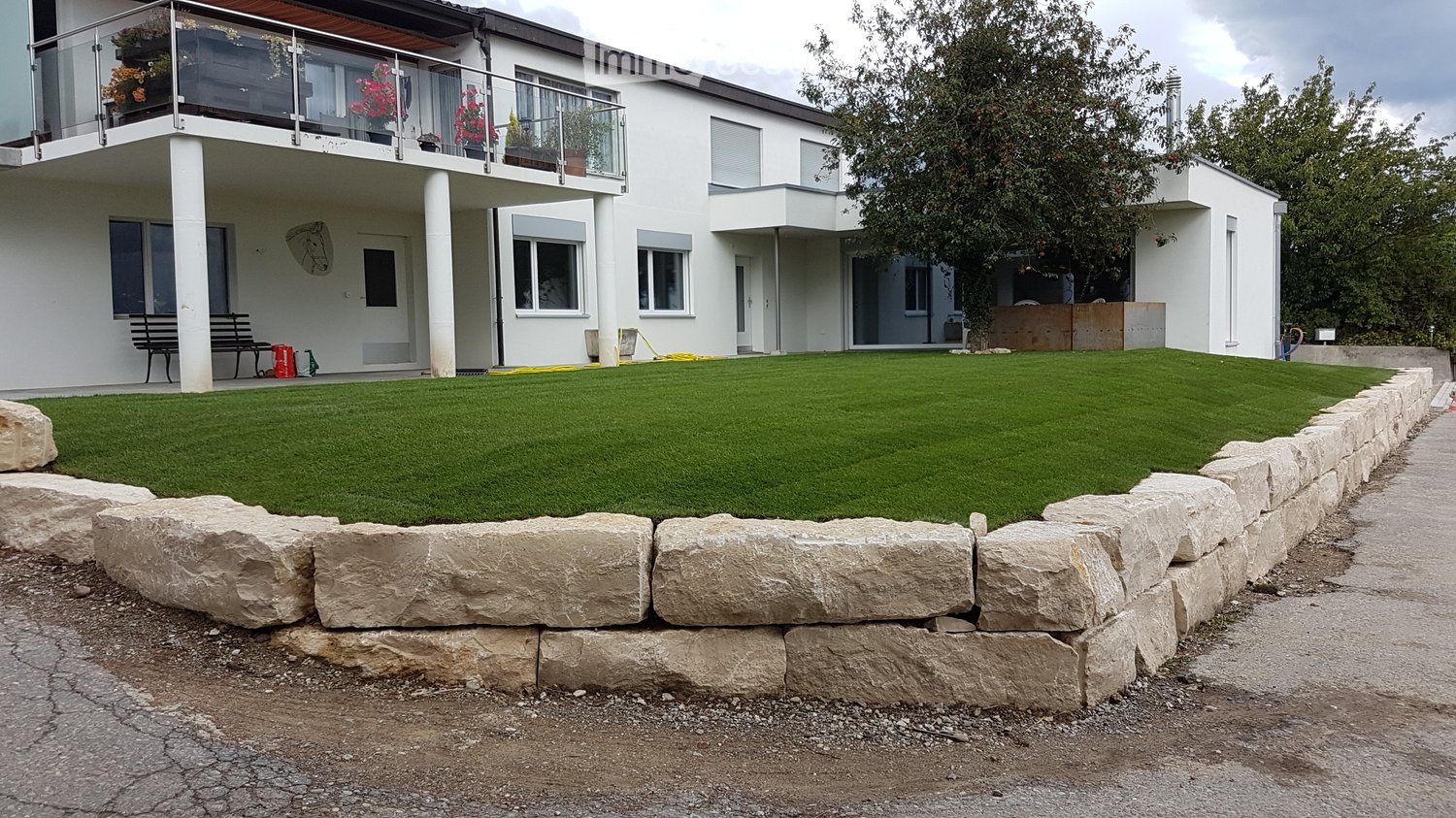 Two-story house, white exterior, balcony with glass railing, lush lawn, stone retaining wall, tree
