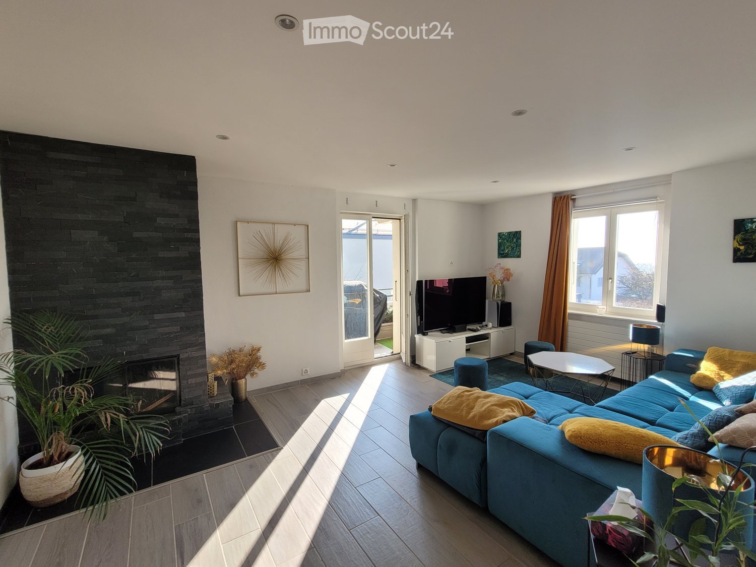 Large room with a blue couch and a black fireplace with a plant on the side. The floor is gray and the wall is white with a window that overlooks the street.