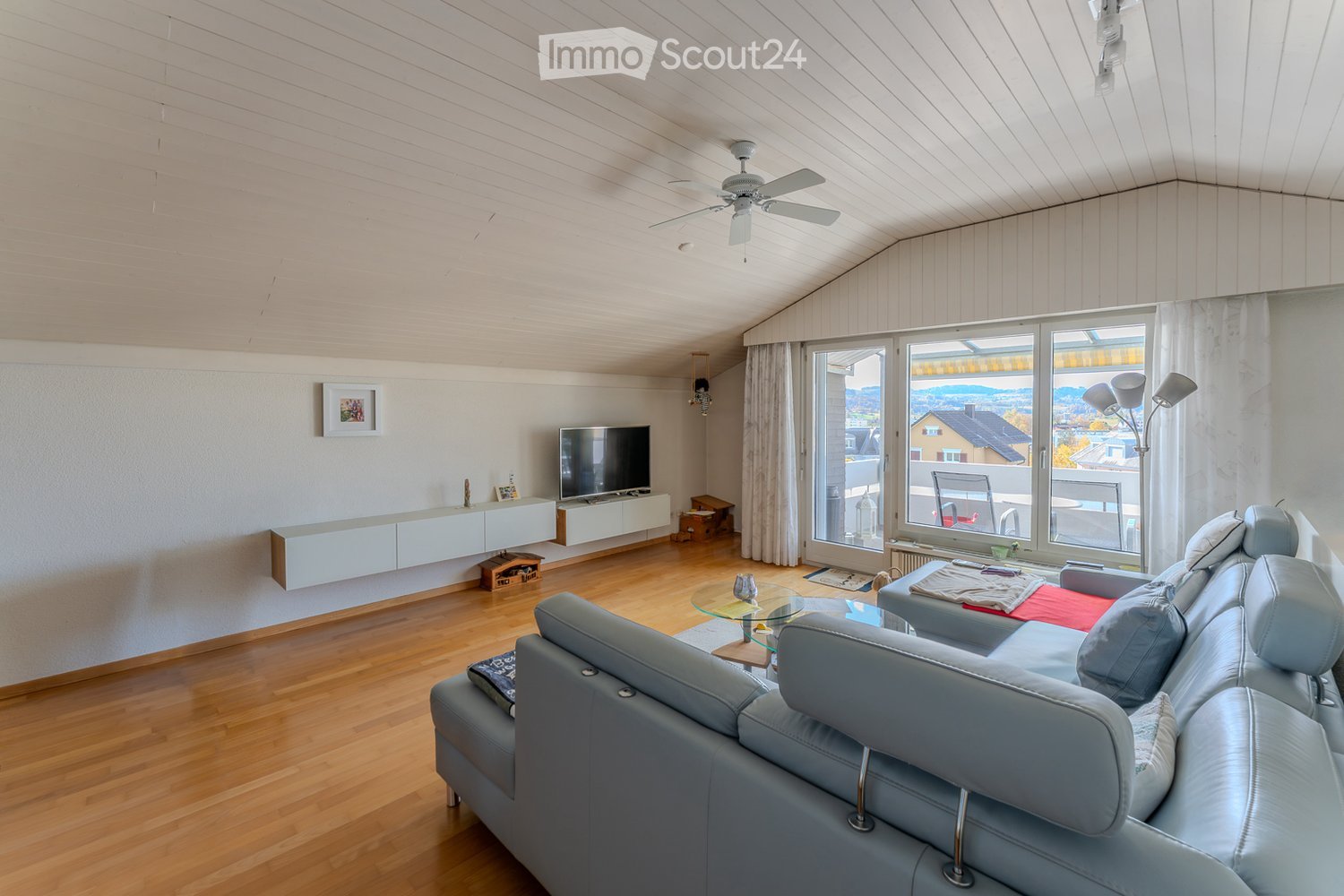 large living area, light wood floors, ceiling fan, large windows, balcony, leather sofa, TV, coffee table, white kitchen cabinets, picture frame