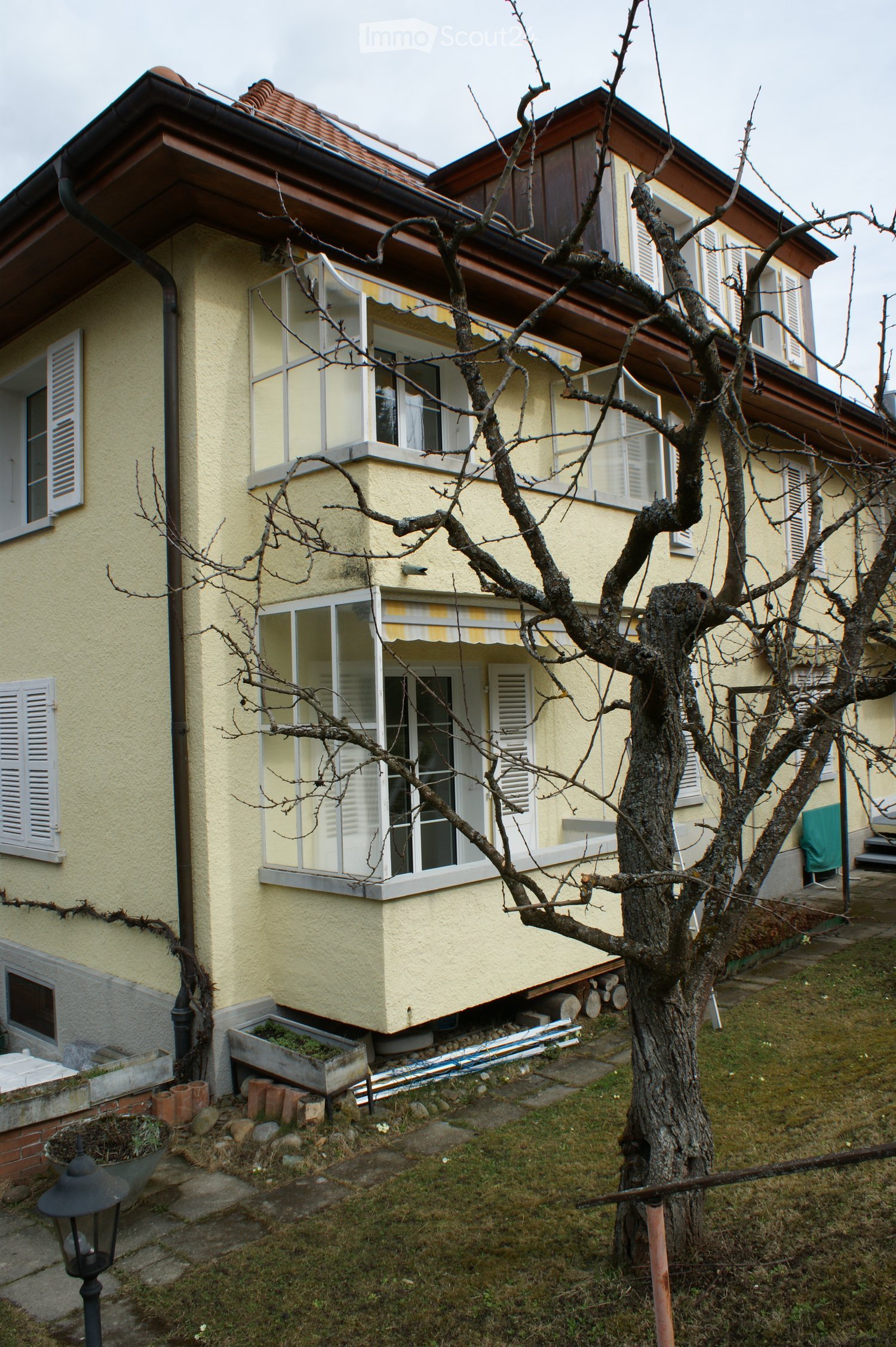 Two-story house, yellow exterior, white shutters, balconies with awnings, slanted roof, garden area with trees
