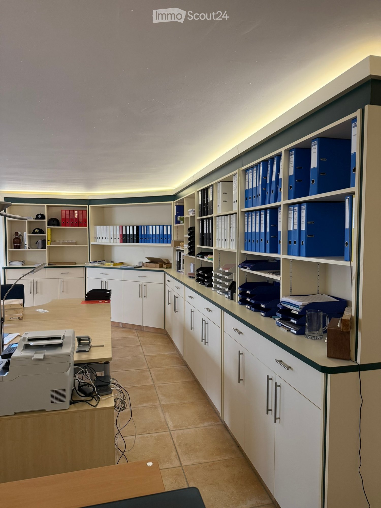 An office space with wooden floors, white cabinets, shelves with folders, desks, and a printer.