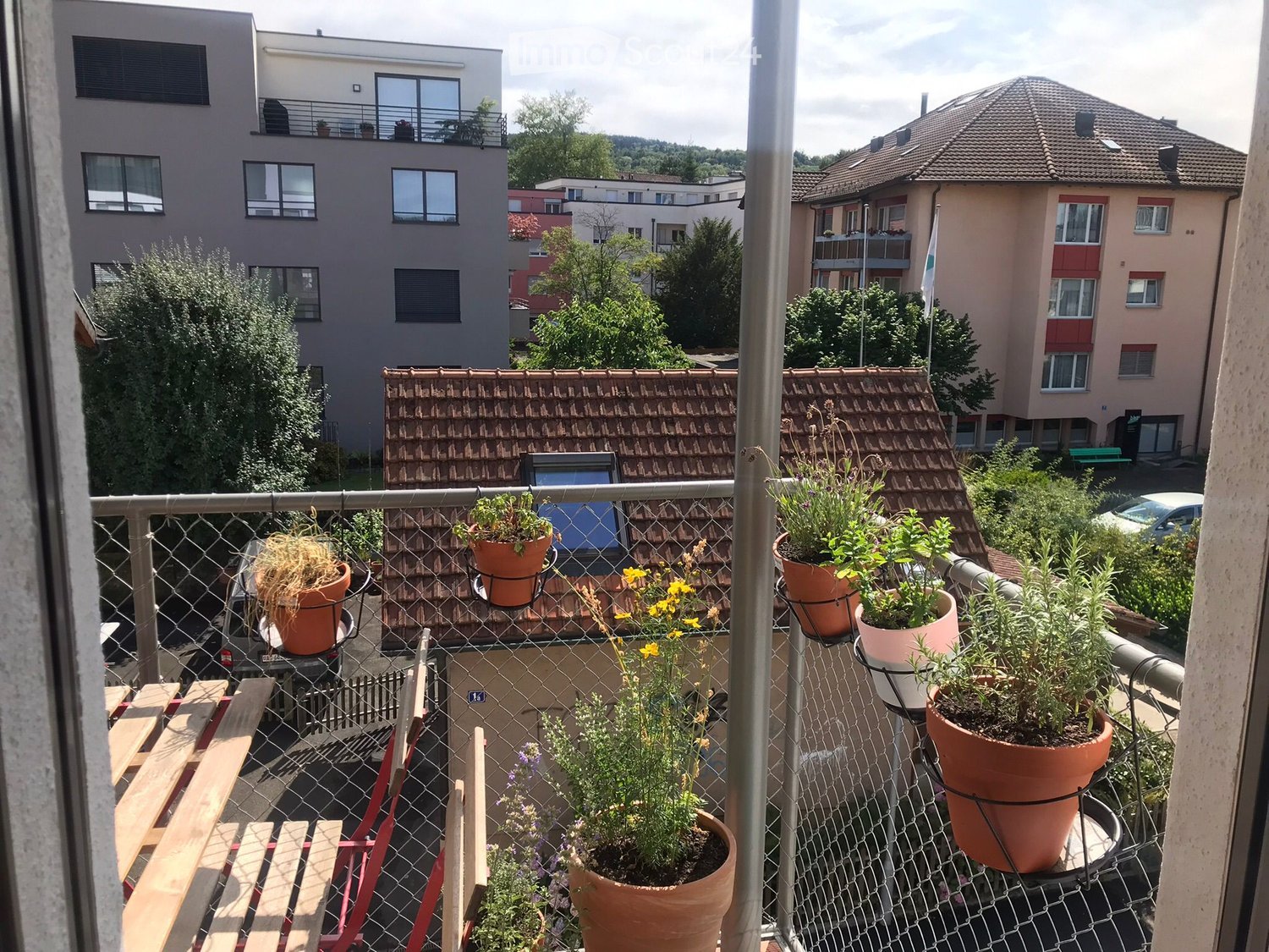 Balcony with plants, metal fence, two wooden chairs, small wooden table, and cars in the distance.