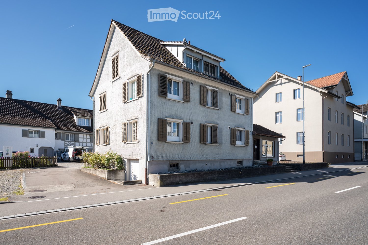 Three-story house with wooden-shuttered windows, red-tiled roof, two parked cars and trees in front.