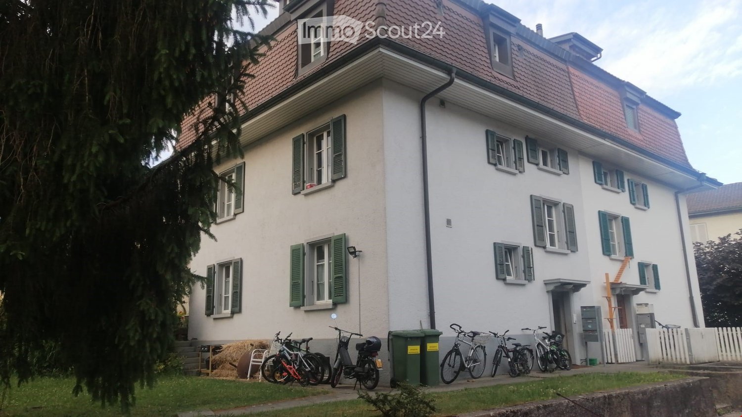 white 3-storey building, red roof, many windows, balconies, green shutters, bikes parked in front
