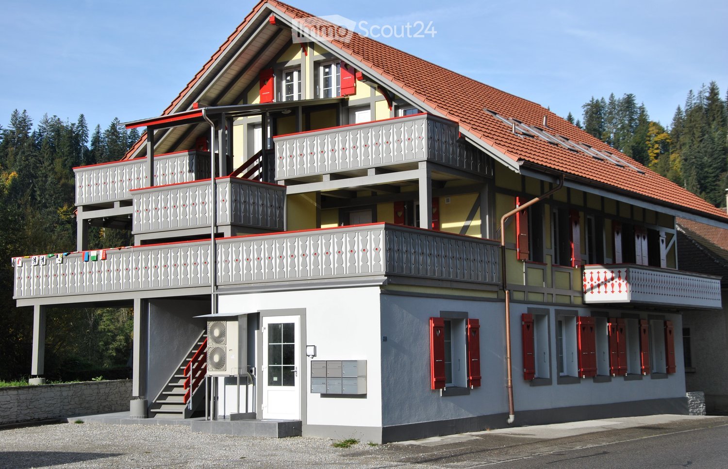 2-story house with red roofs and white walls, balconies, red shutters, exterior air conditioning unit, staircase, mailboxes, gravel driveway, greenery in the background